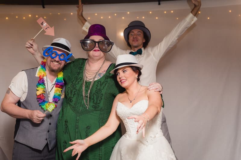 At Kwila Lodge reception, the bride Holly in a white wedding dress and white fedora hat poses with three guests wearing playful accessories including hats, oversized glasses, and a colorful lei.