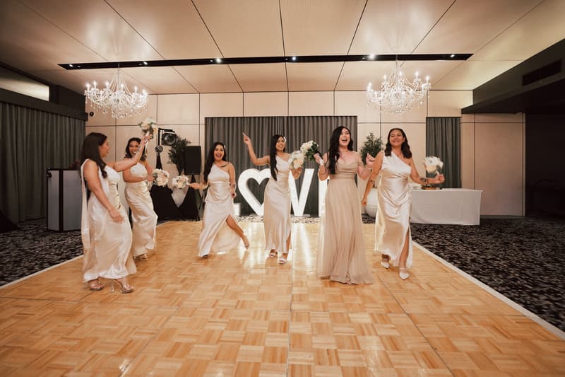 Bridesmaids in matching champagne satin dresses hold bouquets and walk on the dance floor in The Pumicestone Room at Sandstone Point Hotel, with illuminated LOVE letters in the background.