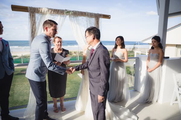 Jason shakes hands with an older man on the ceremony stage at Bilinga Beach Weddings — The Terrace, while Wing stands nearby in her wedding gown. Two bridesmaids in light grey dresses and a woman holding a booklet stand in the background with the ocean visible beyond the railing.