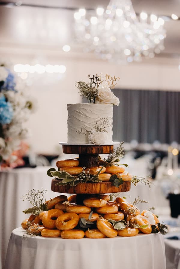 A wedding cake and donut tower decorated with flowers and greenery on a round table at the Sandstone Point Hotel — Pumicestone Room reception.