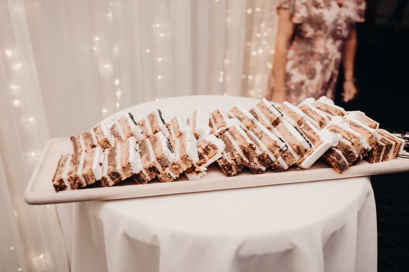 A wooden tray with multiple slices of layered cake arranged on a round table covered with a white tablecloth at The Tides — The Pandanus Room reception.