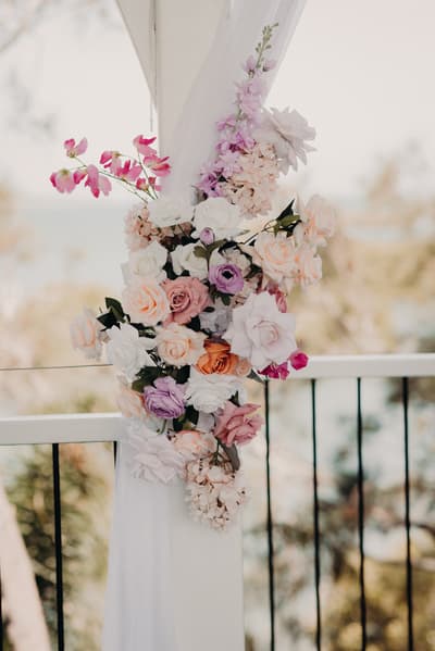 Floral arrangement of pastel roses and other flowers attached to a white draped pillar. Wedding photographer and videographer by She Said Yes.