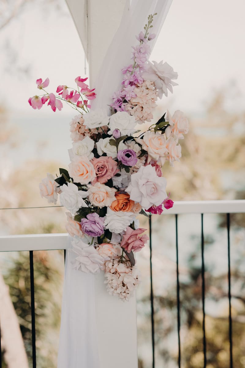Floral arrangement of pastel roses and other flowers attached to a white draped pillar. Destination wedding photographer Australia by She Said Yes.