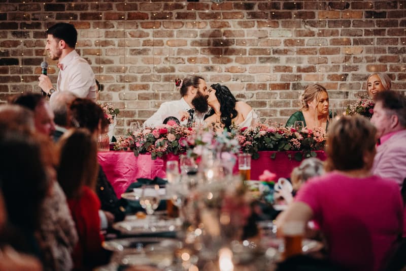 At the Sandstone Point Hotel — Cellar, the bride Mindy and groom David share a kiss at the head table decorated with pink flowers during the wedding reception. A man in a white shirt and bow tie is giving a speech with a microphone to the left, while guests are seated at tables in the foreground.