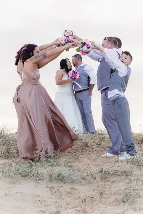 Kelly and Shane kiss while standing on sand with bridesmaids and groomsmen holding bouquets overhead forming an arch at Novotel Sunshine Coast Resort.
