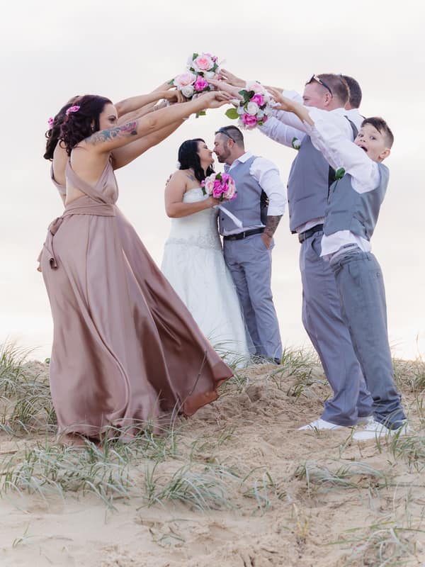 Kelly and Shane kiss while standing on sand with bridesmaids and groomsmen holding bouquets overhead forming an arch at Novotel Sunshine Coast Resort.