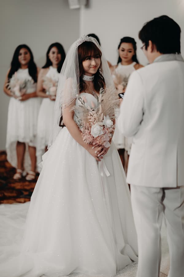 The bride Henny stands holding a bouquet and facing the groom Timothy, who is dressed in a white suit, during their wedding ceremony at Royal on the Park. Bridesmaids in white dresses holding bouquets stand in the background.