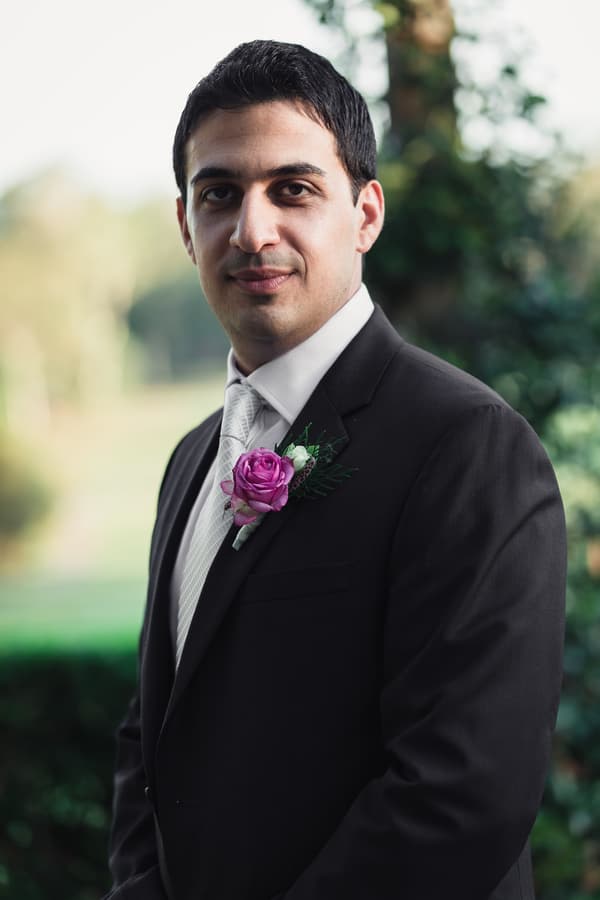The groom Pasha stands outdoors with a blurred natural background, wearing a dark suit, white shirt, white tie, and a purple boutonniere.