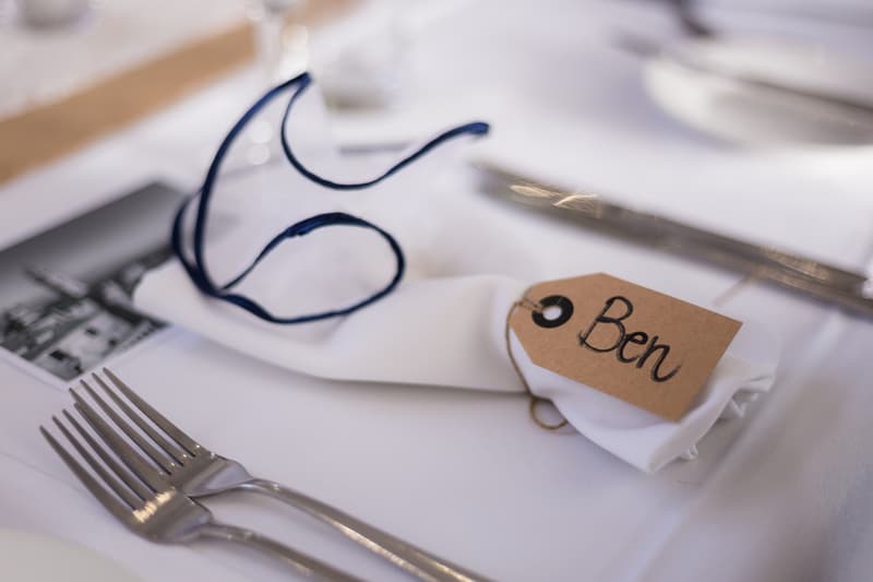 Place setting at the reception table in The Malouf Room at Toowong Rowing Club with a white napkin tied with a navy blue ribbon and a name tag labeled 'Ben'.