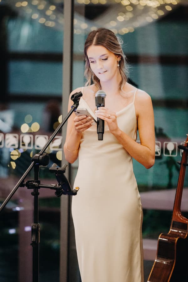 The bride Libby stands at a microphone reading from her phone during the reception at The Tides — The Pandanus Room.