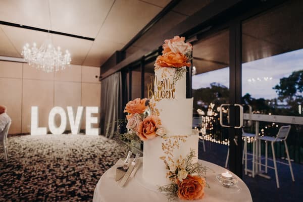 Three-tier white wedding cake decorated with orange and white flowers and gold accents on a round table at Sandstone Point Hotel — Pumicestone Room, with illuminated LOVE letters and a chandelier in the background.