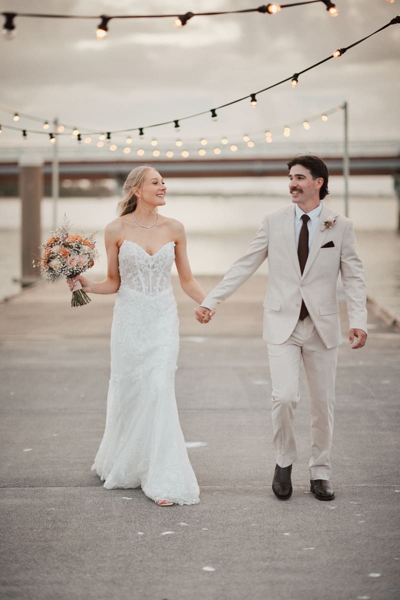 Morgan and Joel walk hand in hand outdoors, with Morgan holding a bouquet and string lights overhead. Getting ready photos by She Said Yes.