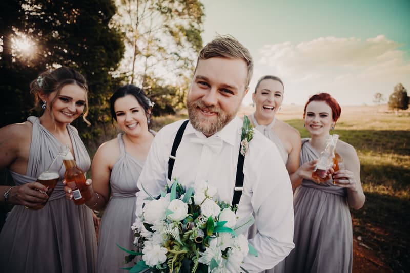 The groom Trent stands holding a bouquet of white flowers with four bridesmaids behind him, each wearing matching grey dresses and holding bottles of drinks, at Ocean View Estates outdoors.
