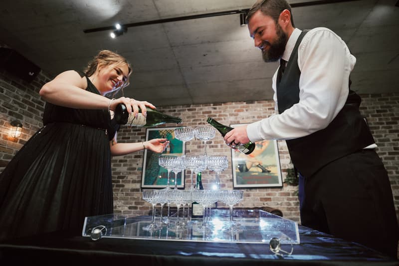 The bride Jayme and the groom Christopher pour champagne into a stacked pyramid of glasses at the reception stage of Sandstone Point Hotel — The Cellar.