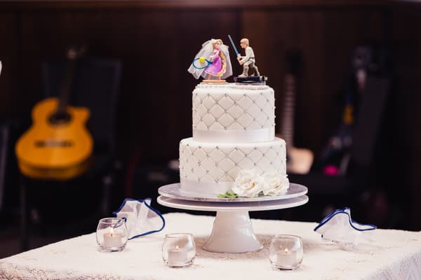 Two-tier white wedding cake with quilted pattern and pearl accents, topped with bride and groom figurines, displayed on a white cake stand at Toowong Rowing Club — The Malouf Room. The cake table is decorated with white flowers and surrounded by four small lit candles in glass holders. Background shows blurred guitars and dark wood paneling.