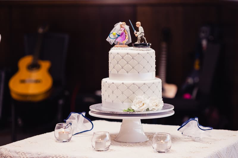 Two-tier white wedding cake with quilted pattern and pearl accents, topped with bride and groom figurines, displayed on a white cake stand at Toowong Rowing Club — The Malouf Room. The cake table is decorated with white flowers and surrounded by four small lit candles in glass holders. Background shows blurred guitars and dark wood paneling.