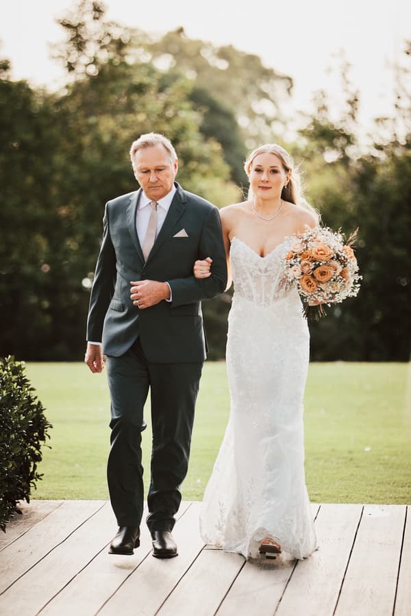 The bride Morgan walks arm-in-arm with an older man, likely her father, along a wooden pathway at Sandstone Point Hotel — The Pavilion, holding a bouquet of flowers.