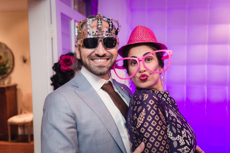 A man in a light gray suit and tie wearing a jeweled crown and sunglasses poses next to a woman in a dark lace dress wearing oversized pink glasses and a pink hat at Hillstone St Lucia — The Rosewood Room.