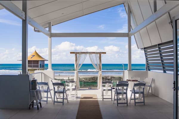 Empty ceremony stage at Bilinga Beach Weddings — The Terrace with white chairs arranged facing a wooden arch draped with sheer fabric, overlooking the ocean and a lifeguard tower.
