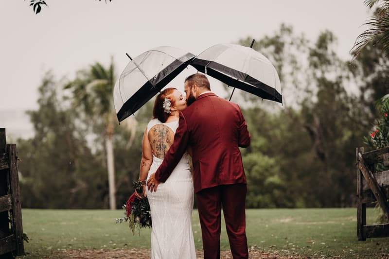 The bride and groom stand outdoors under clear umbrellas, facing away from the camera, with the bride holding a bouquet and the groom wearing a red suit jacket.