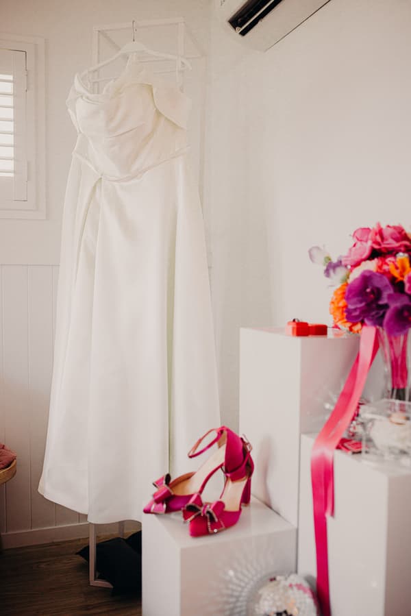 The bride's white wedding dress hangs on a stand at Sandstone Point Hotel, with a pair of pink high-heeled shoes and a bouquet of colorful flowers placed nearby on white pedestals.