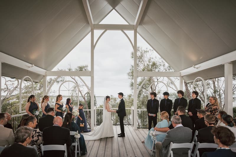 Terion and Tyler stand facing each other holding hands at the altar during their wedding ceremony at Sandstone Point Hotel — The Pavilion, surrounded by bridesmaids, groomsmen, a flower girl, and seated guests.