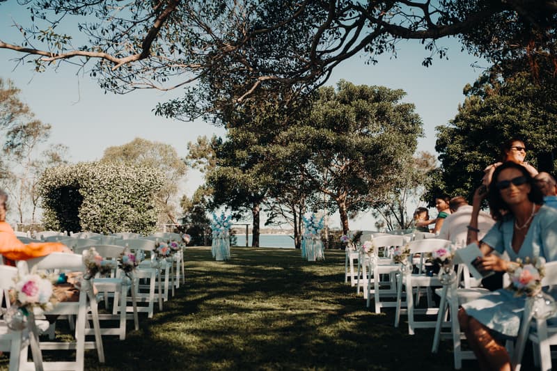 Guests seated on white chairs arranged in rows at the outdoor ceremony stage of Sandstone Point Hotel — The Pavilion, with floral decorations on the chairs and a view of trees and water in the background.
