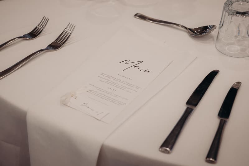 Close-up of a white tablecloth with a printed menu card, two forks, two knives, a spoon, and an upside-down glass at The Tides — The Pandanus Room reception.