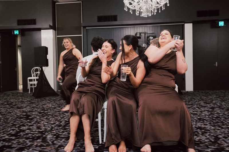 Three bridesmaids in brown dresses sit on chairs back-to-back with a man in a white shirt at Sandstone Point Hotel — The Pumicestone Room, holding glasses of water and laughing, while another bridesmaid walks barefoot in the background holding a microphone.