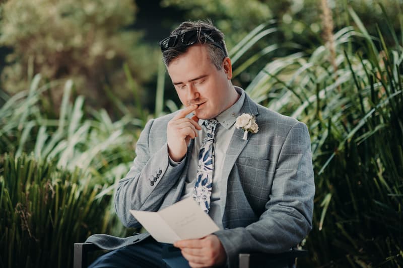 The groom Dale sits outdoors at Sandstone Point Hotel reading a card, wearing a grey checked suit, floral tie, and sunglasses on his head.
