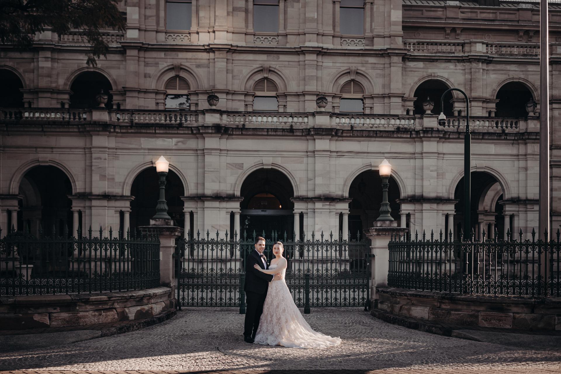 The bride Aria and groom Antony stand together in front of the Queensland Parliament building, posing for a couple portrait with the building's ornate stone facade and wrought iron fence behind them.