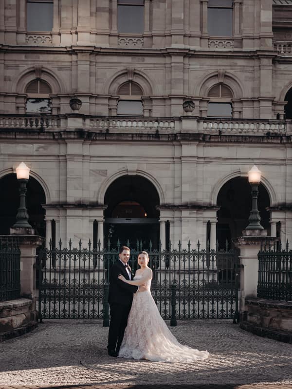 The bride Aria and groom Antony stand together in front of the Queensland Parliament building, posing for a couple portrait with the building's ornate stone facade and wrought iron fence behind them.