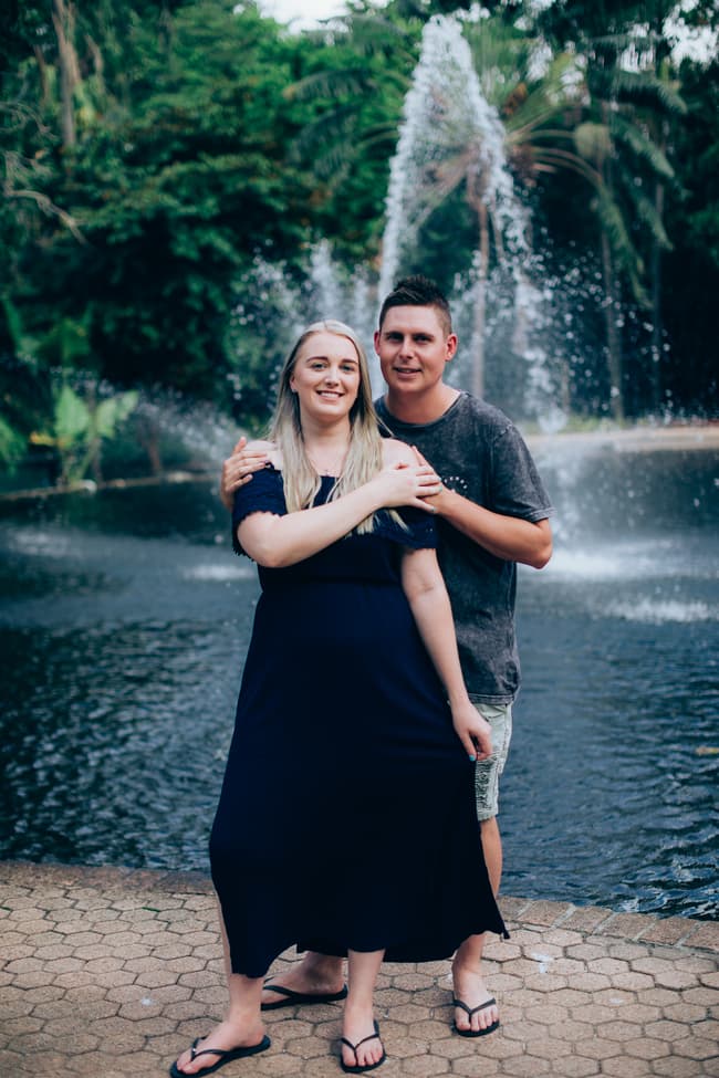 The couple standing close together, smiling by a fountain at City Botanic Gardens.