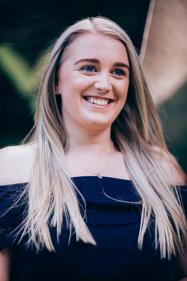 A person smiling brightly, with long hair and a necklace, at City Botanic Gardens.