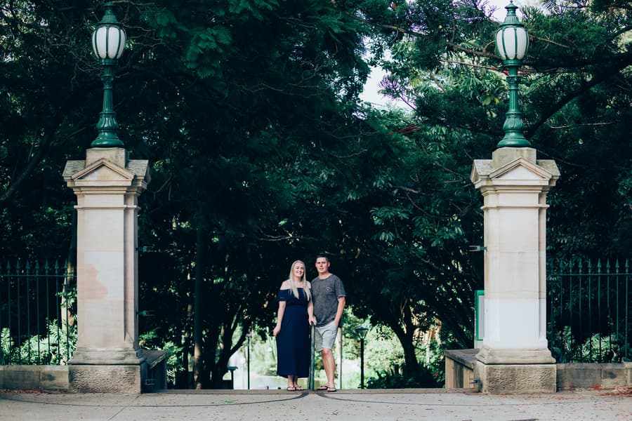 The couple standing between two stone pillars with vintage lamps at City Botanic Gardens, surrounded by lush greenery.