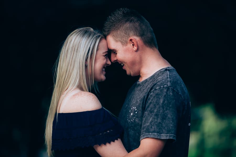 The couple standing close, foreheads touching, smiling warmly at each other in a shaded area.