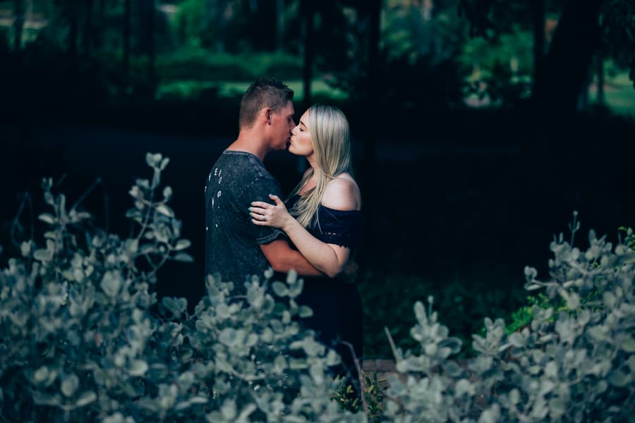 The couple sharing a kiss surrounded by greenery, with soft focus on foliage in the foreground.