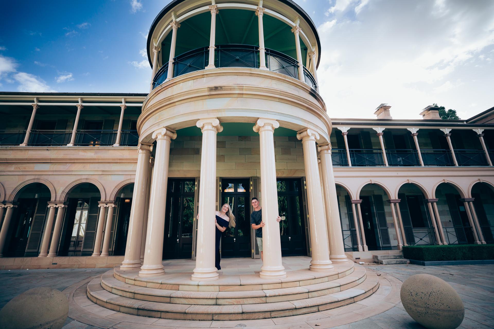The couple peeking from behind columns of Old Government House, playful and joyful.