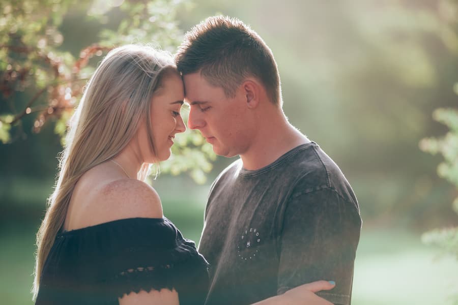 The couple standing close with foreheads touching, bathed in soft sunlight filtering through trees.