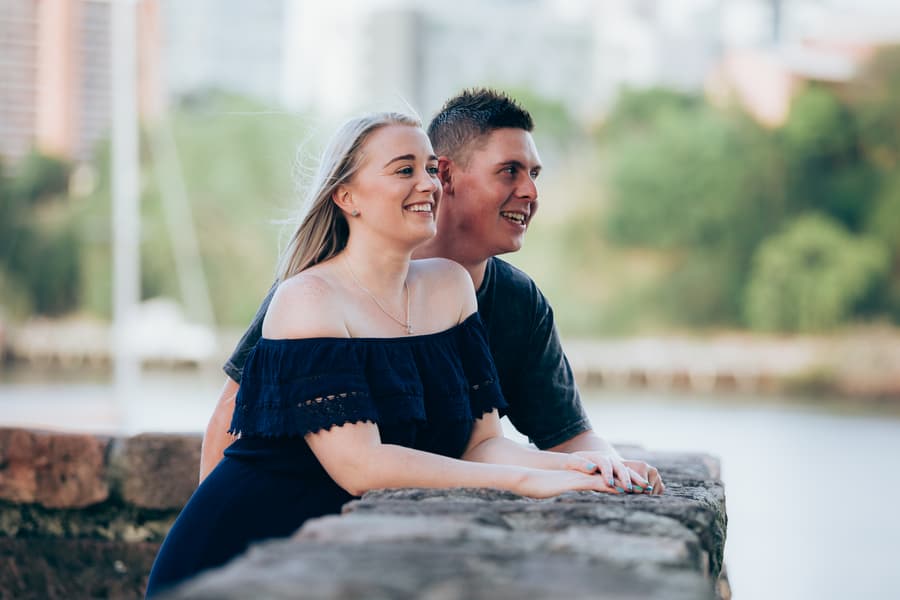 The couple leaning on a stone wall, smiling and looking into the distance with a cityscape backdrop.