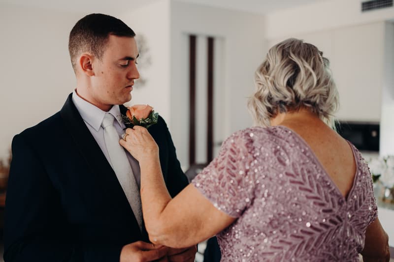 The groom is being helped with his suit jacket by a man in a white shirt in a room with a brown leather couch and a wall picture of a wooden staircase outdoors.