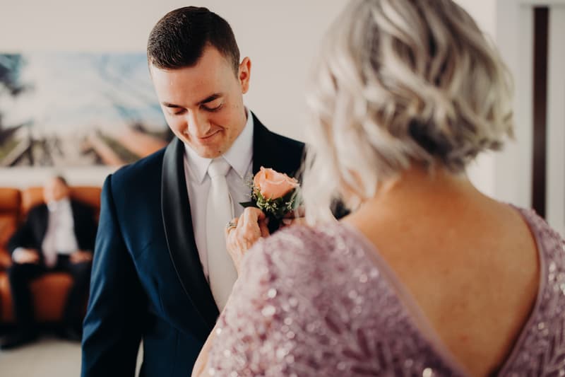 The groom Dylan stands while an older woman, likely the mother of the groom, pins a peach rose boutonniere on his navy blue tuxedo jacket at Sandstone Point Hotel.