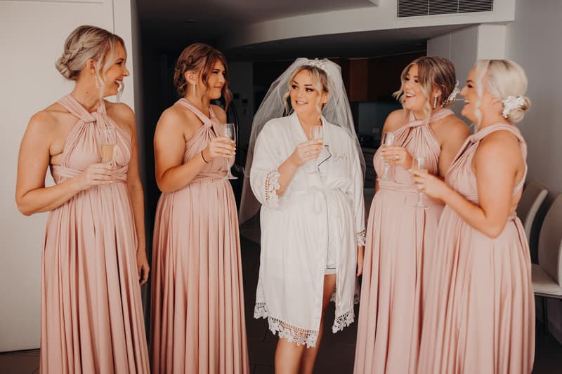 The bride Emily in a white robe and veil toasts with four bridesmaids dressed in matching blush pink gowns indoors at Sandstone Point Hotel.