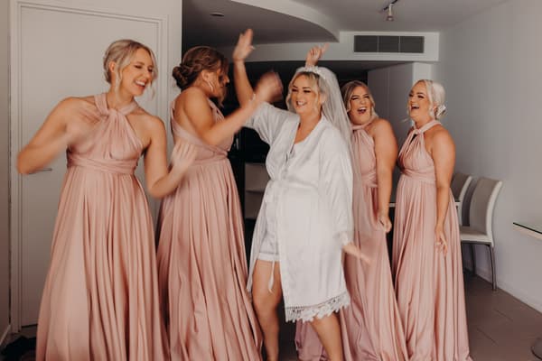 Two wedding rings displayed in a small round wooden box with a cushioned interior, placed on a reflective surface with blurred fabric and greenery in the background.