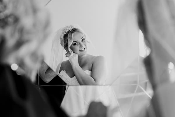 Emily, the bride, poses indoors wearing a strapless white wedding gown and veil, holding a bouquet of mixed flowers including roses and greenery.