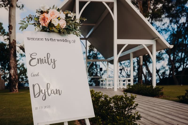 The bride pins a boutonniere on an older man, likely the father of the bride, while holding a young boy dressed in a white shirt and bow tie.