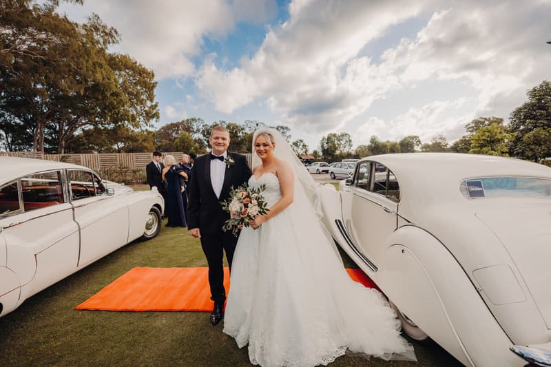 Emily the bride and Dylan the groom sit inside a vintage white car, with Emily holding a bouquet of flowers.