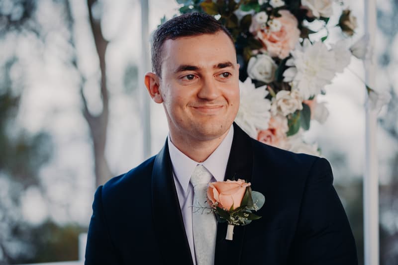 The bride Emily, wearing a white wedding gown and veil, is walking arm-in-arm with an older man in a black tuxedo, likely her father, on the lawn at Sandstone Point Hotel — Pavilion. Emily holds a bouquet of flowers as they approach the ceremony area.