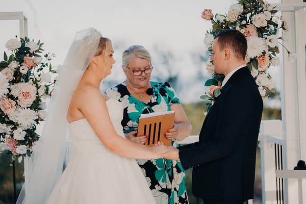 Emily and Dylan stand facing each other at the ceremony stage at Sandstone Point Hotel — Pavilion, surrounded by bridesmaids in blush pink dresses holding bouquets on the left and groomsmen in black suits with pink ties on the right. An officiant stands behind the bride.