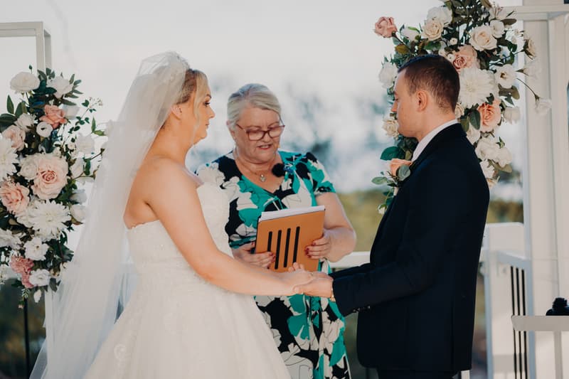 Emily and Dylan stand facing each other at the ceremony stage at Sandstone Point Hotel — Pavilion, surrounded by bridesmaids in blush pink dresses holding bouquets on the left and groomsmen in black suits with pink ties on the right. An officiant stands behind the bride.
